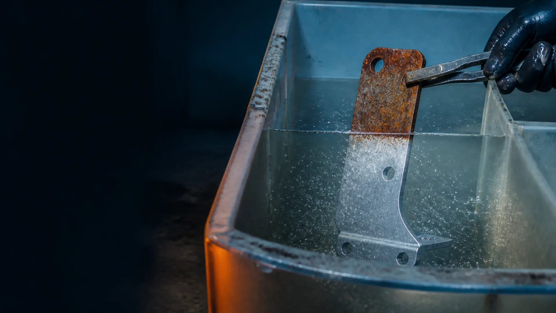 Rusted steel bracket half-submerged in an acidic cleaning bath, the submerged half bright