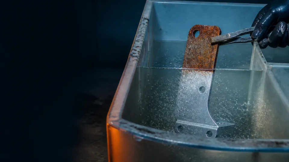 Rusted steel bracket half-submerged in an acidic cleaning bath, the submerged half bright